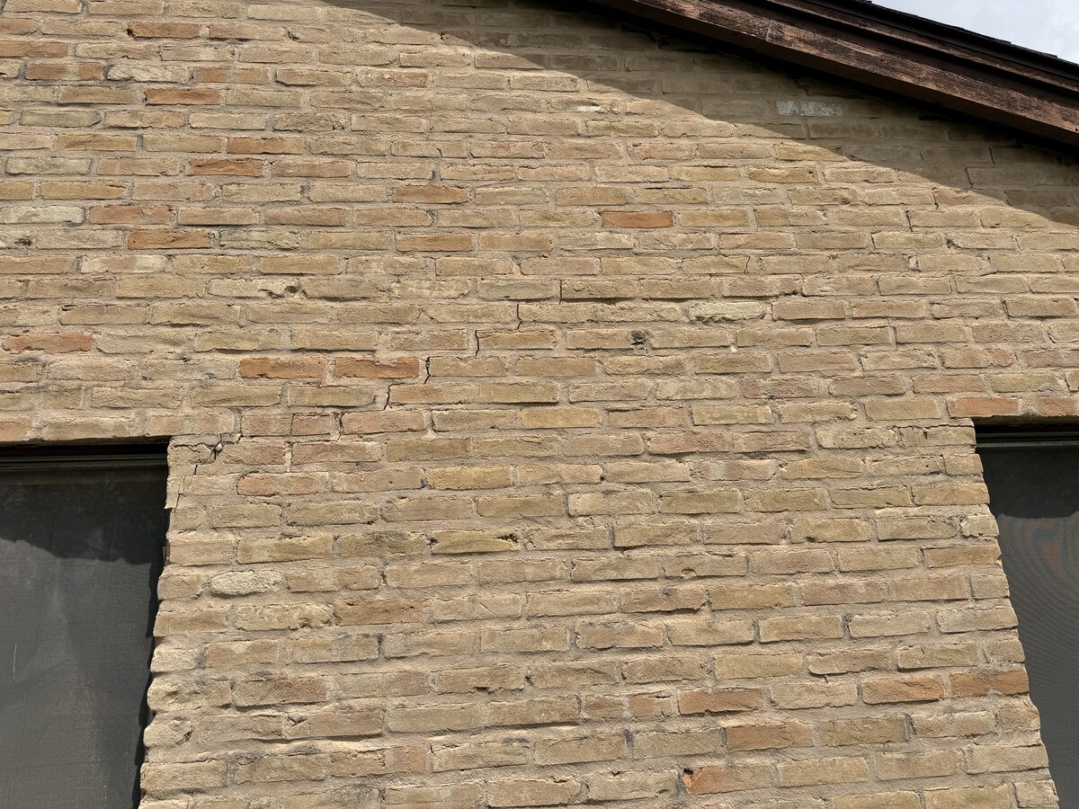 Network of settlement cracks spread across a brick gable wall above the windows of a one-story home