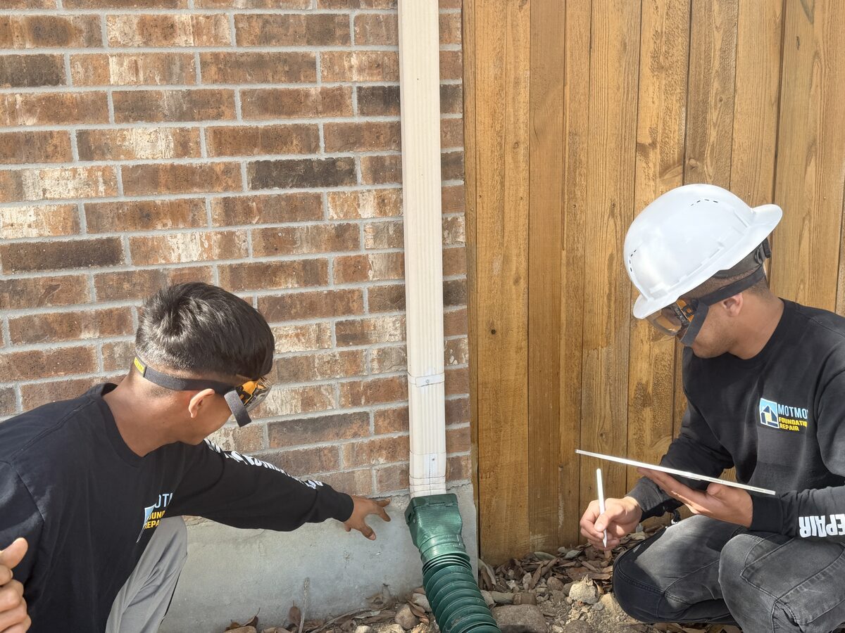 Crew members inspecting an exposed slab edge and downspout drainage at a brick home's foundation