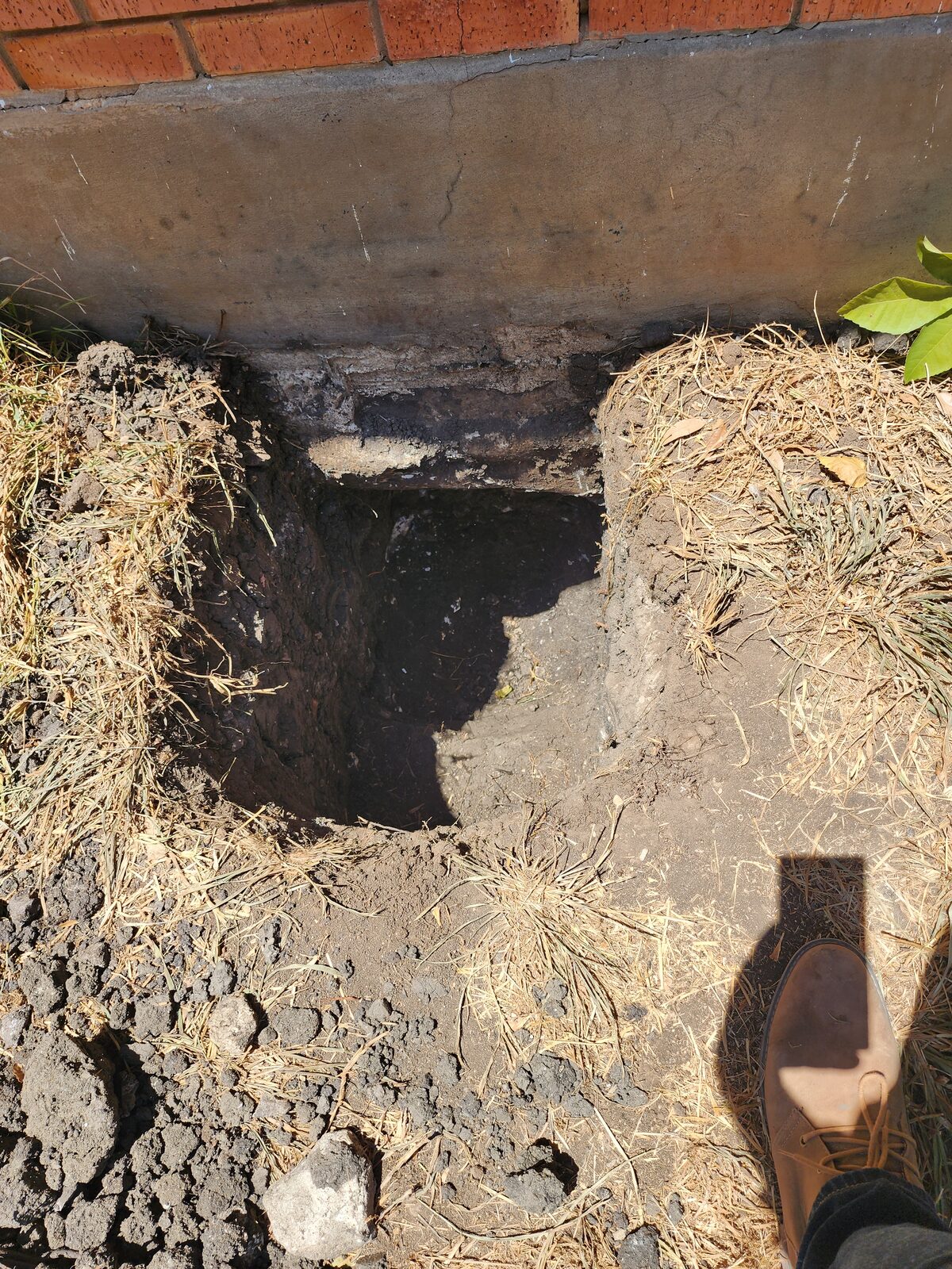 Hand-dug pit exposing the grade beam beneath a brick home's foundation, ready for pier installation
