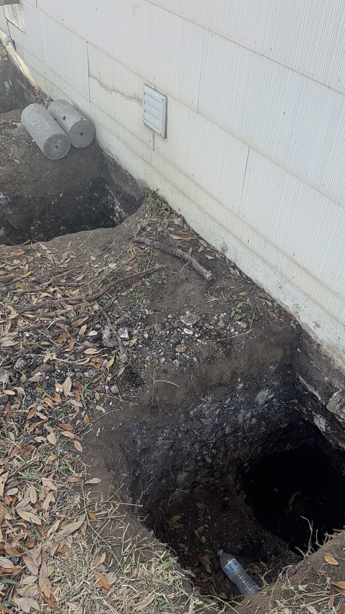 Pier hole excavated at the base of a siding-clad wall to access the foundation for repair