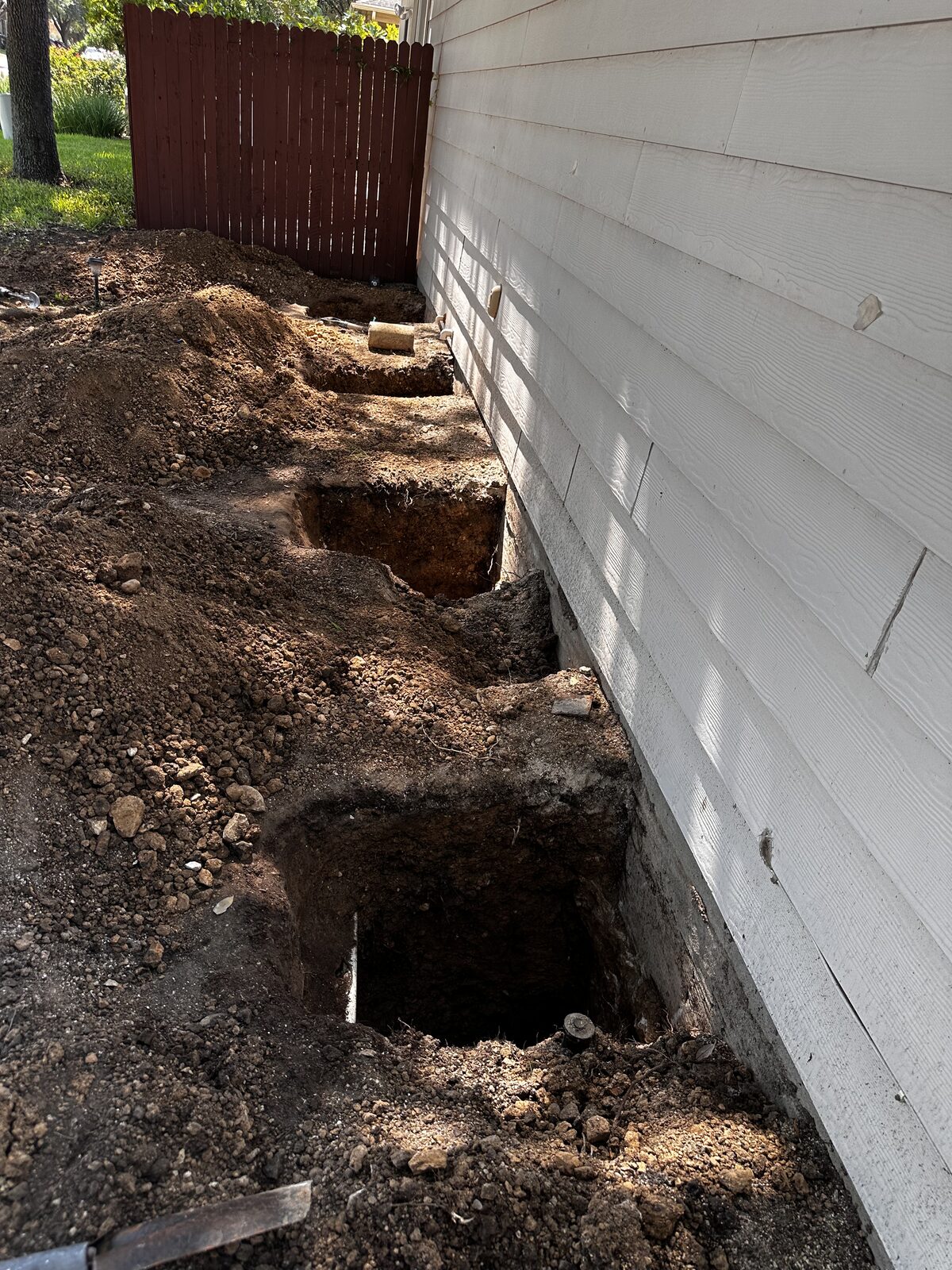 Hand-dug pier holes spaced along the slab edge of a home in preparation for foundation piers