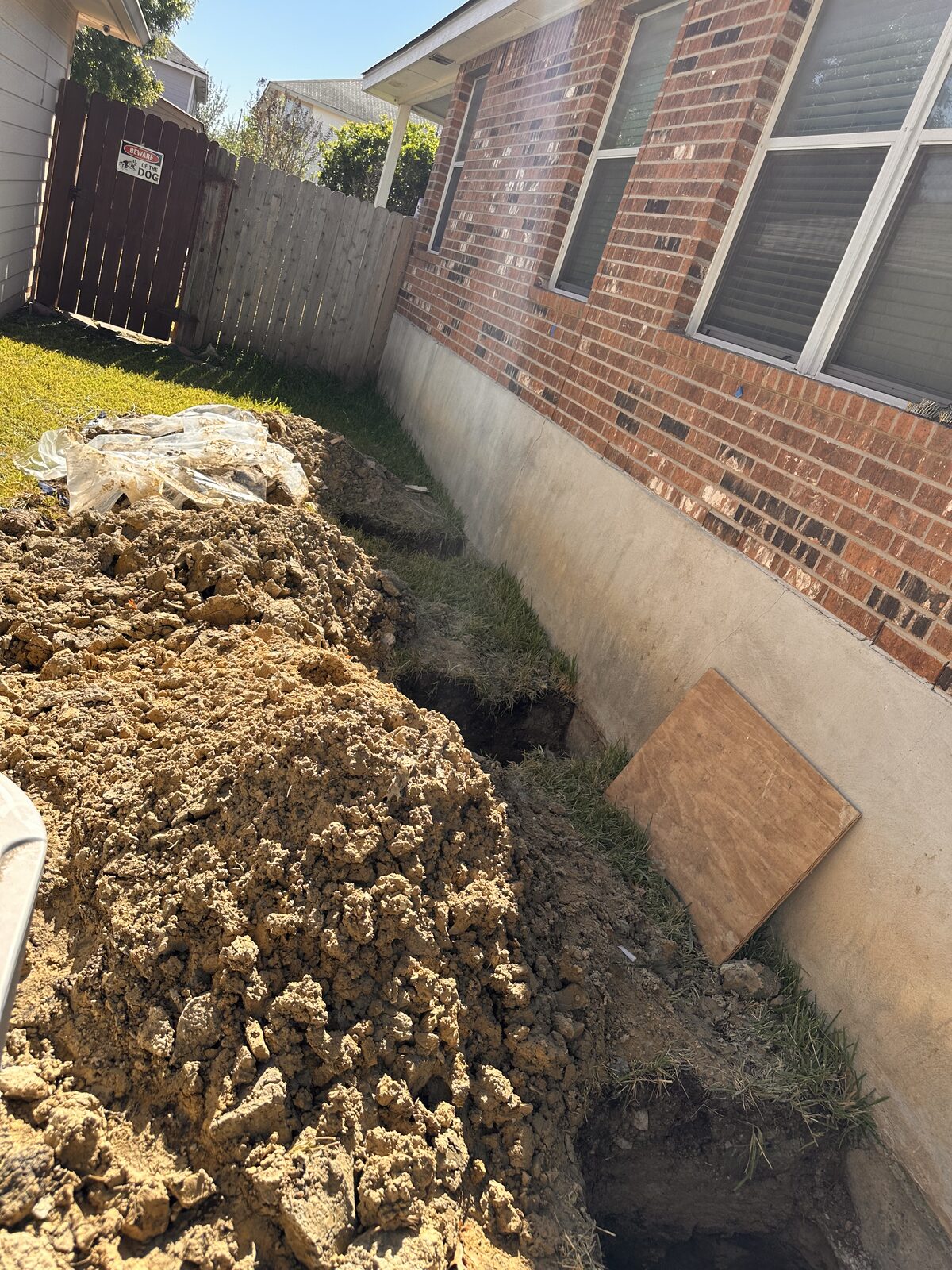 Side-yard view of foundation excavation along a brick home with a large spoil mound and plywood covers