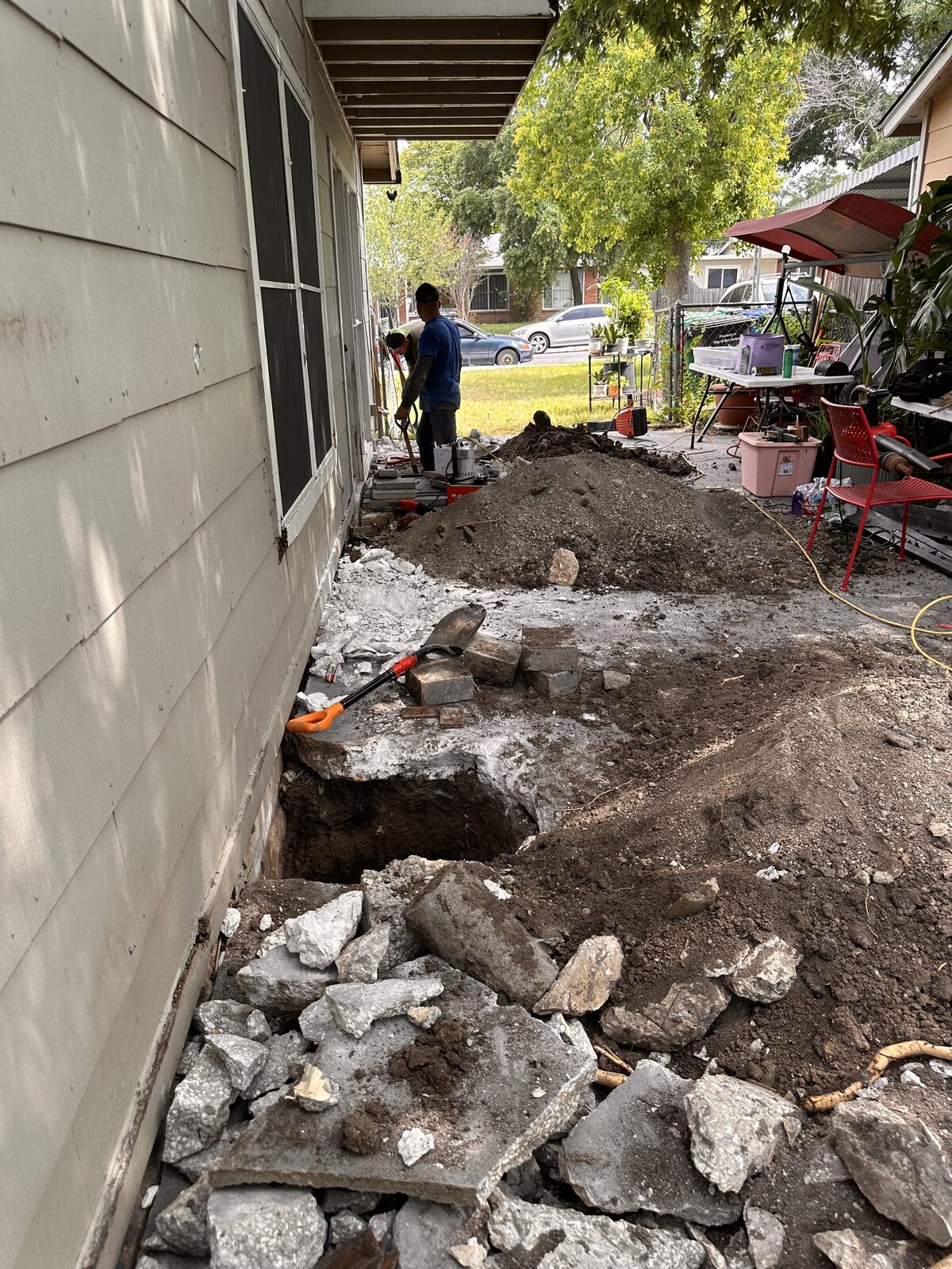 Crew hand-digging pier holes along a home's foundation in a narrow side yard, rock spoil piled beside the wall