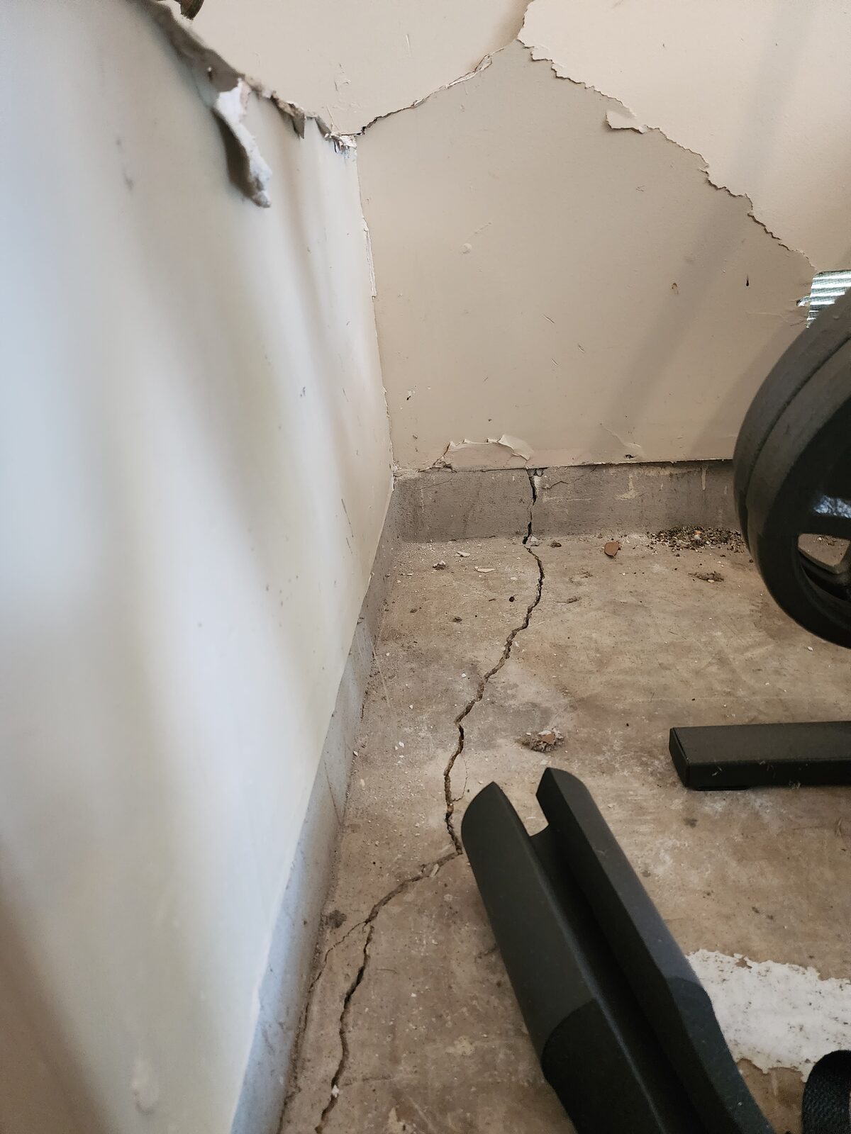 Crack running across a concrete garage slab into a damaged drywall corner, evidence of foundation movement