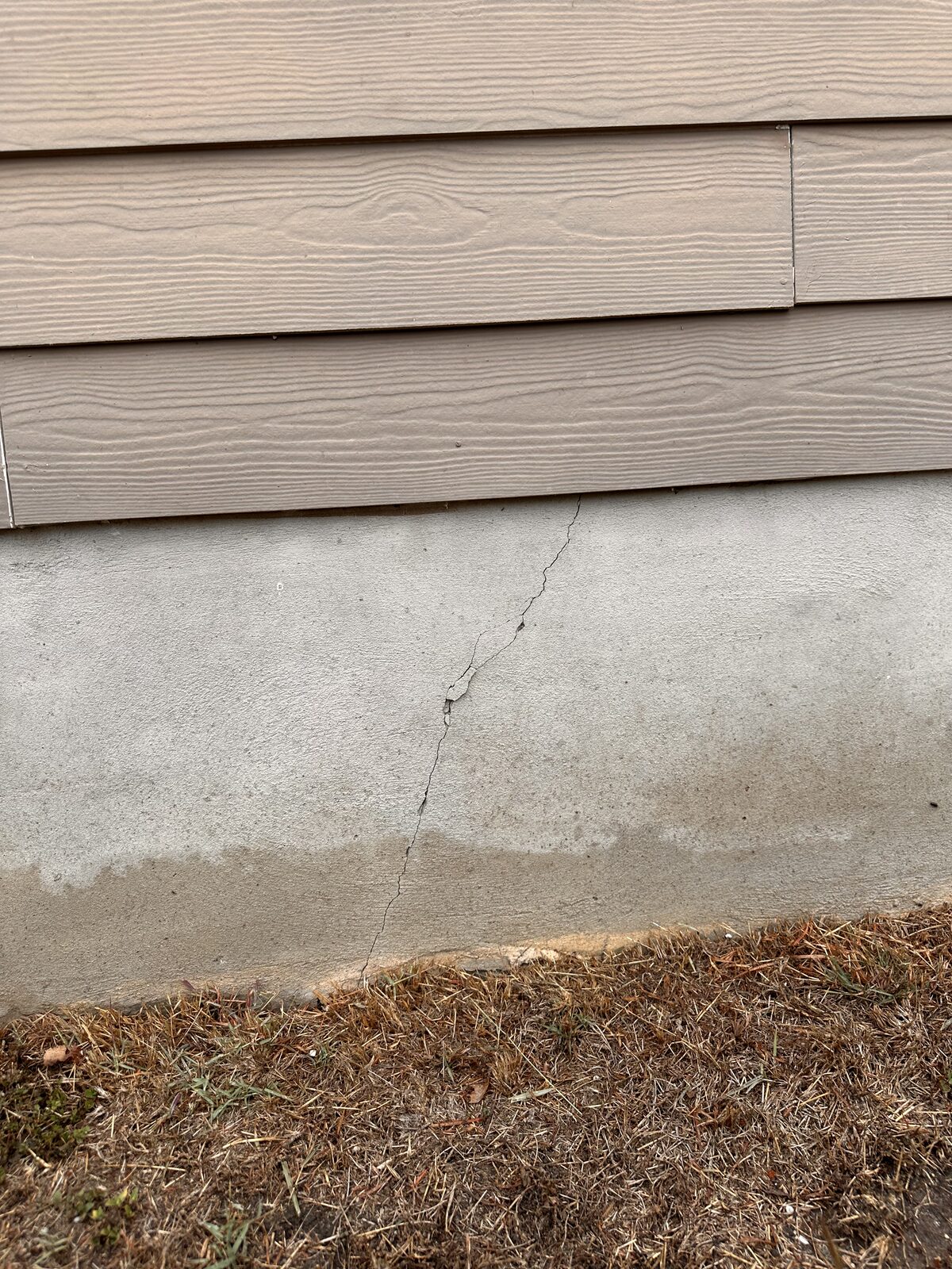 Hairline crack in an exposed concrete slab edge beneath lap siding, an early sign of foundation settlement