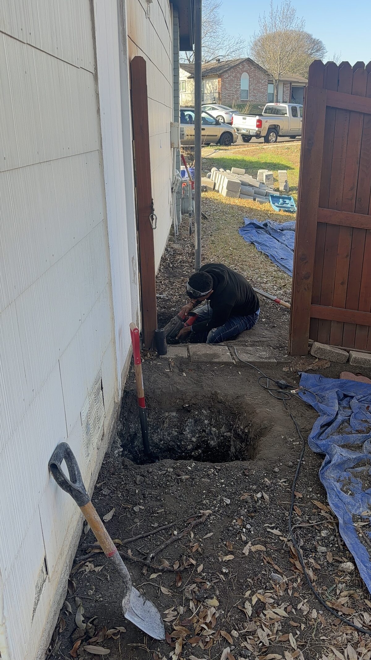 Crew member hand-digging a foundation pier hole beside a home with a shovel and protective tarp