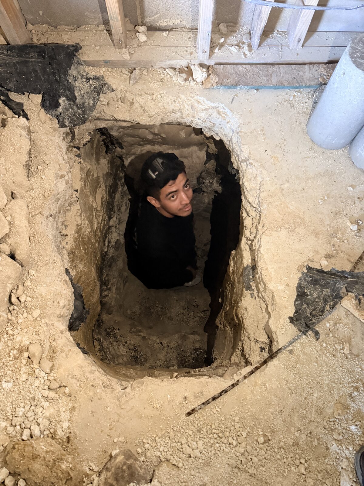 Foundation repair technician standing in a hand-dug pier pit beneath a slab, looking up from the excavation