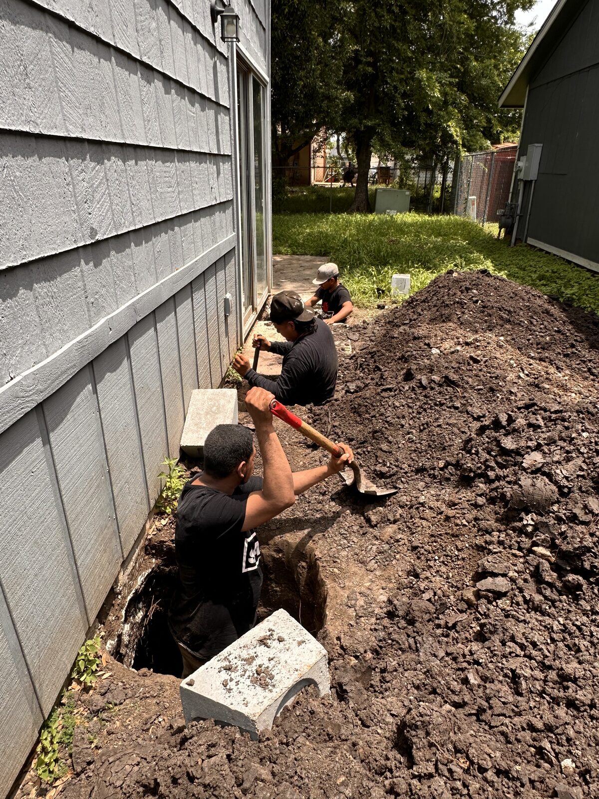 Crew member breaking soil with a mattock in a pier pit, with concrete pile cylinders staged along the house
